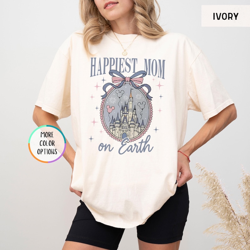 May include: Ivory-colored t-shirt with the text "Happiest Mom on Earth" above a castle graphic. The graphic includes a bow, stars, and Mickey Mouse balloons. The shirt is a relaxed fit.