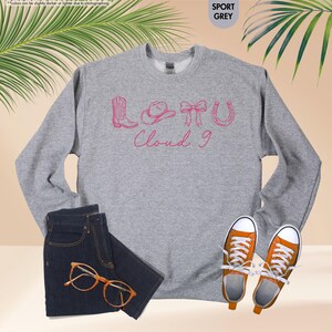 May include: A grey sweatshirt featuring a pink graphic of a cowboy boot, hat, bow, and horseshoe, with the text "Cloud 9". The image also shows a pair of dark blue jeans, orange-framed glasses, and orange trainers.
