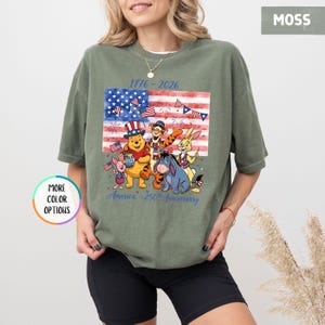 May include: Moss green t-shirt featuring a graphic of the American flag with Winnie the Pooh and friends. The design includes the text "1776-2026" and "America - 250th Anniversary". The shirt has a relaxed fit.