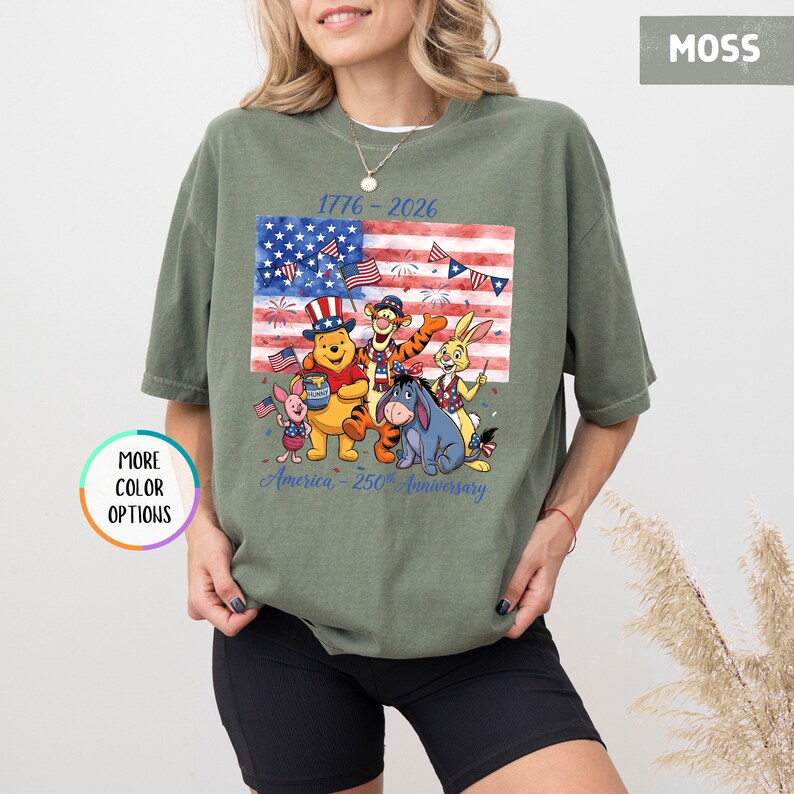 May include: Moss green t-shirt featuring a graphic of the American flag with Winnie the Pooh and friends. The design includes the text "1776-2026" and "America - 250th Anniversary". The shirt has a relaxed fit.