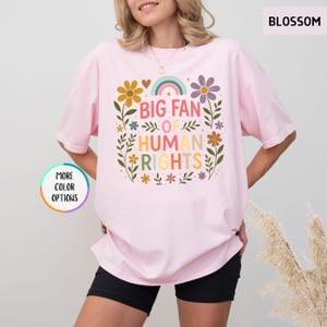 May include: A light pink t-shirt with the text "BIG FAN OF HUMAN RIGHTS" in colorful letters. The design includes flowers, a rainbow, and leaves. The shirt is a relaxed fit.
