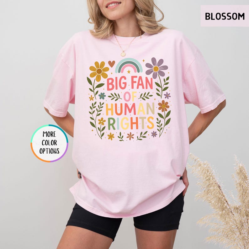 May include: A light pink t-shirt with the text "BIG FAN OF HUMAN RIGHTS" in colorful letters. The design includes flowers, a rainbow, and leaves. The shirt is a relaxed fit.
