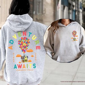 Puede incluir: Una sudadera con capucha gris claro con un gr&aacute;fico colorido de una casa atada a globos, con las palabras "ADVENTURE AWAITS PARADISE FALLS" y "SINCE 2009". La sudadera tiene un bolsillo delantero y una capucha. El texto "SPORT GREY" est&aacute; en la esquina superior izquierda.