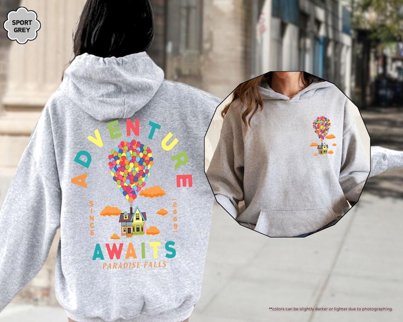 Puede incluir: Una sudadera con capucha gris claro con un gr&aacute;fico colorido de una casa atada a globos, con las palabras "ADVENTURE AWAITS PARADISE FALLS" y "SINCE 2009". La sudadera tiene un bolsillo delantero y una capucha. El texto "SPORT GREY" est&aacute; en la esquina superior izquierda.