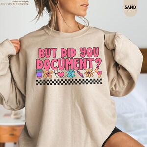 Puede incluir: Sudadera color arena con el texto rosa "BUT DID YOU DOCUMENT?". El dise&ntilde;o incluye iconos m&eacute;dicos como una taza de caf&eacute;, una venda, un estetoscopio y una jeringa, con un borde a cuadros blanco y negro.