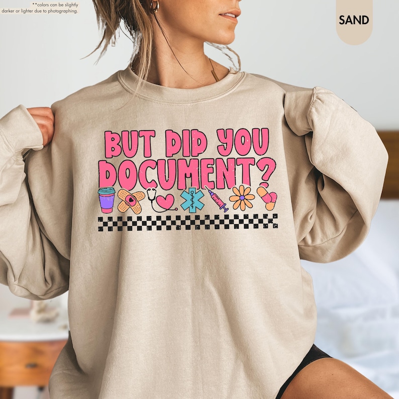Puede incluir: Sudadera color arena con el texto rosa "BUT DID YOU DOCUMENT?". El dise&ntilde;o incluye iconos m&eacute;dicos como una taza de caf&eacute;, una venda, un estetoscopio y una jeringa, con un borde a cuadros blanco y negro.