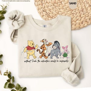 Puede incluir: Una sudadera de color arena con un gr&aacute;fico de Winnie the Pooh, Tigger, Eeyore y Piglet. El texto "without Pooh, the adventure would be impossible" est&aacute; impreso debajo. La sudadera tiene mangas largas.