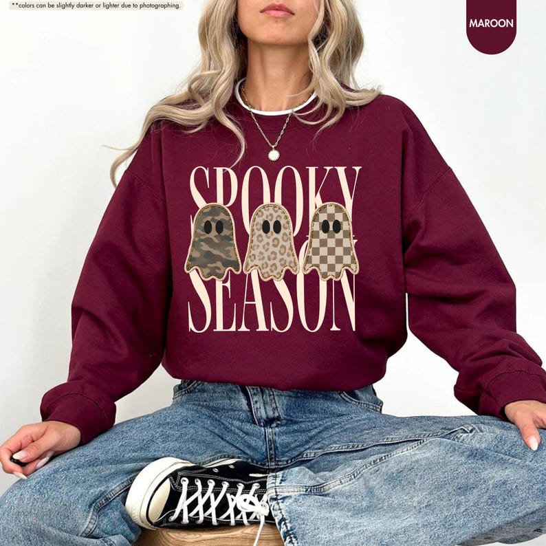 May include: A maroon crewneck sweatshirt featuring the text SPOOKY SEASON in a cream-colored serif font. Three ghost illustrations are centered between the words, each with a different pattern: camouflage, leopard print, and a brown checkerboard.