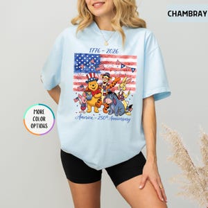 May include: Light blue t-shirt featuring a graphic of the American flag with cartoon characters. The shirt has the text "1776-2026" and "America - 250th Anniversary". The shirt has a round neck and short sleeves.