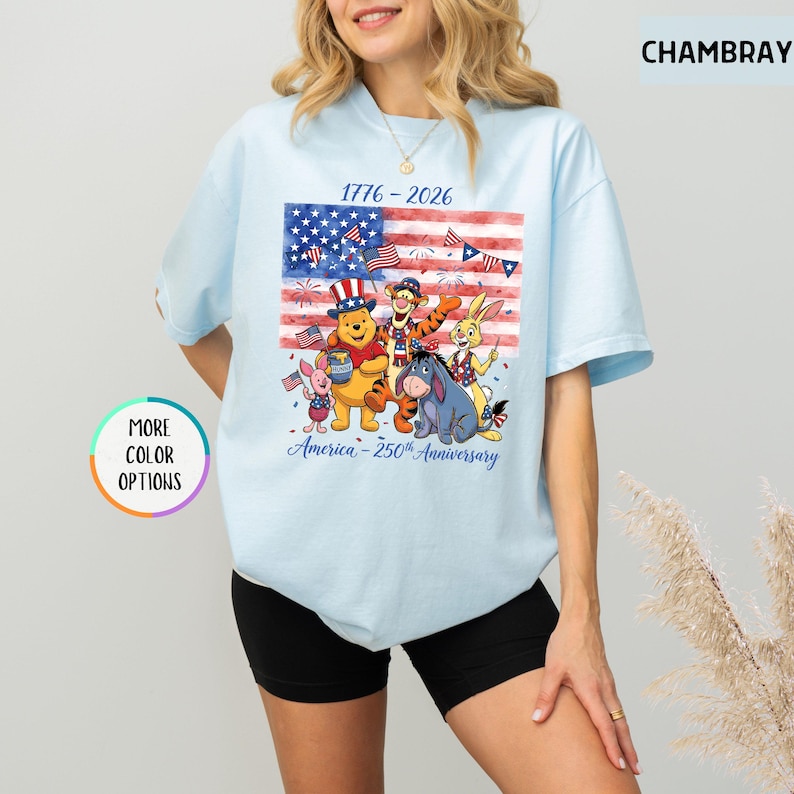 May include: Light blue t-shirt featuring a graphic of the American flag with cartoon characters. The shirt has the text "1776-2026" and "America - 250th Anniversary". The shirt has a round neck and short sleeves.