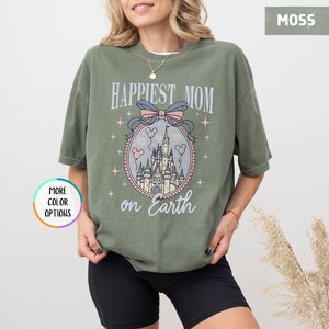May include: Moss green t-shirt with the text "Happiest Mom on Earth" and a castle graphic. The graphic is in a circular frame with a pink bow and stars. The shirt is a relaxed fit.