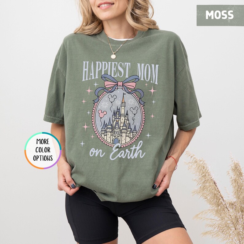 May include: Moss green t-shirt with the text "Happiest Mom on Earth" and a castle graphic. The graphic is in a circular frame with a pink bow and stars. The shirt is a relaxed fit.