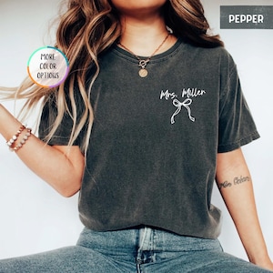 Puede incluir: Una camiseta gris oscuro con el texto "Mrs. Miller" y un dise&ntilde;o de lazo blanco. La camiseta tiene cuello redondo y mangas cortas. La imagen tambi&eacute;n incluye un bot&oacute;n "More Color Options" y la palabra "Pepper".