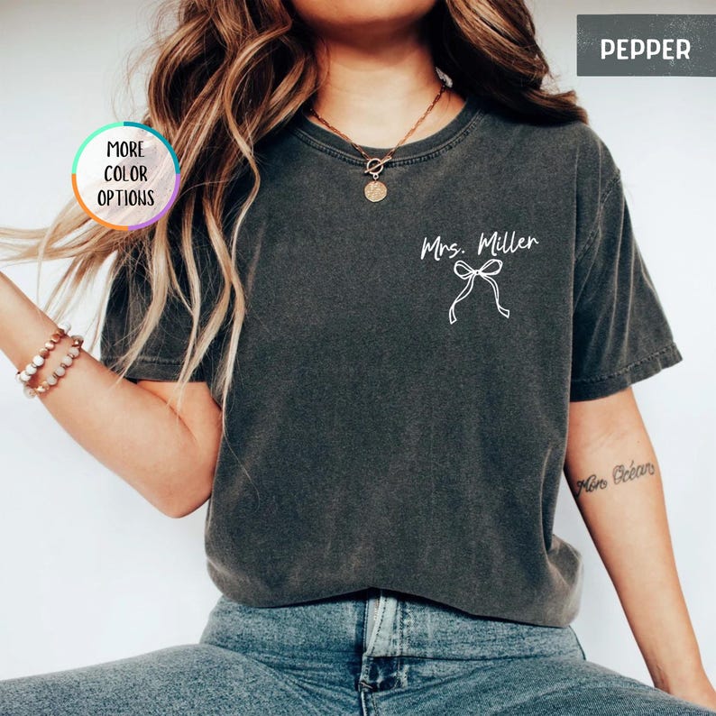 Puede incluir: Una camiseta gris oscuro con el texto "Mrs. Miller" y un dise&ntilde;o de lazo blanco. La camiseta tiene cuello redondo y mangas cortas. La imagen tambi&eacute;n incluye un bot&oacute;n "More Color Options" y la palabra "Pepper".