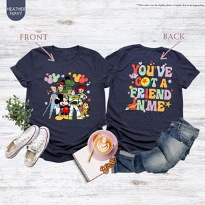 May include: Heather navy blue t-shirts featuring a front graphic of cartoon characters and the text "YOU'VE GOT A FRIEND IN ME" on the back. The front graphic includes Mickey Mouse, Buzz Lightyear, and other characters. The shirts are displayed with jeans and shoes.