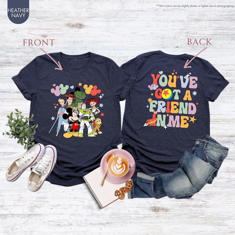 May include: Heather navy blue t-shirts featuring a front graphic of cartoon characters and the text "YOU'VE GOT A FRIEND IN ME" on the back. The front graphic includes Mickey Mouse, Buzz Lightyear, and other characters. The shirts are displayed with jeans and shoes.