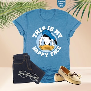 Op de afbeelding: Heather deep teal t-shirt met een afbeelding van Donald Duck en de tekst "THIS IS MY HAPPY FACE". De outfit omvat een donkerblauwe jeans, een bril en cr&egrave;mekleurige mocassins.