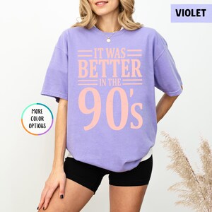 May include: A violet short-sleeved t-shirt featuring the light peach-colored text "IT WAS BETTER IN THE 90's" printed on the front. The shirt has a crew neckline and a relaxed fit, with a small circular graphic on the left side reading "MORE COLOR OPTIONS".