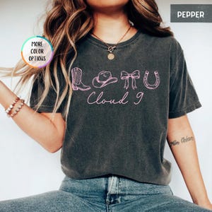 May include: A dark gray t-shirt with a pink graphic featuring a cowboy boot, hat, bow, and horseshoe, with the words "Cloud 9" in a script font. The shirt has a vintage, worn look.