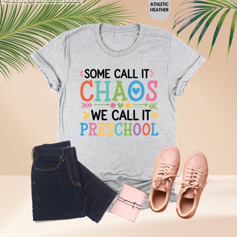 May include: A light gray t-shirt with the text "SOME CALL IT CHAOS WE CALL IT PRESCHOOL" in colorful letters. The shirt is paired with dark blue jeans, pink sneakers, and a small pink wallet.