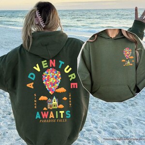 Puede incluir: Una sudadera con capucha verde militar con un gr&aacute;fico colorido en la espalda con el texto "ADVENTURE AWAITS PARADISE FALLS" y un dise&ntilde;o de globo aerost&aacute;tico. La parte delantera de la sudadera con capucha tiene un gr&aacute;fico m&aacute;s peque&ntilde;o de un globo aerost&aacute;tico y una casa. La sudadera con capucha tiene una capucha y un bolsillo delantero.