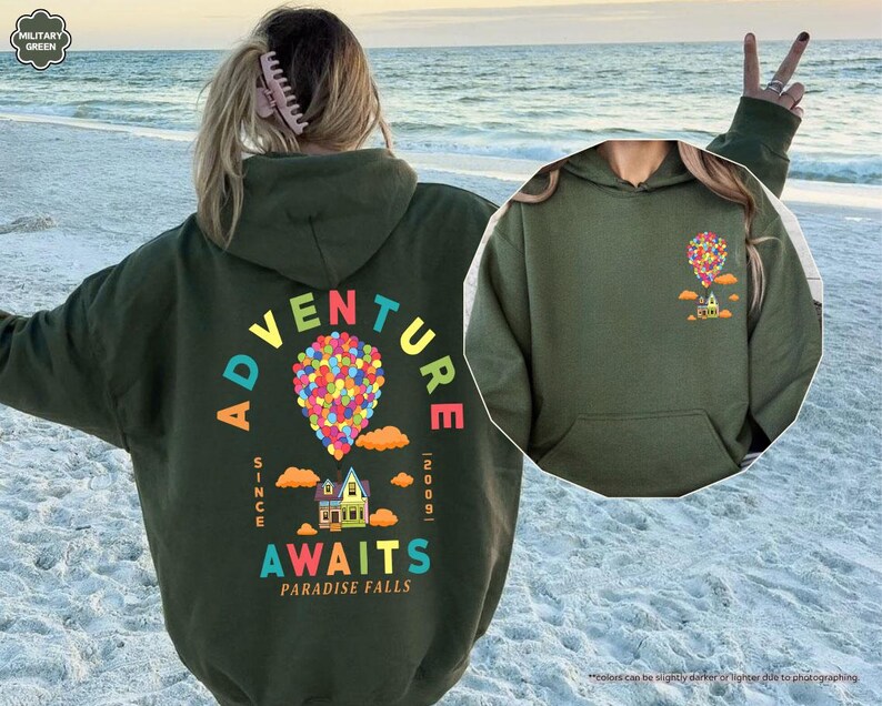 Puede incluir: Una sudadera con capucha verde militar con un gr&aacute;fico colorido en la espalda con el texto "ADVENTURE AWAITS PARADISE FALLS" y un dise&ntilde;o de globo aerost&aacute;tico. La parte delantera de la sudadera con capucha tiene un gr&aacute;fico m&aacute;s peque&ntilde;o de un globo aerost&aacute;tico y una casa. La sudadera con capucha tiene una capucha y un bolsillo delantero.