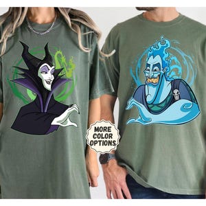 Puede incluir: Dos camisetas verde oliva con personajes de dibujos animados. Una camiseta muestra a Mal&eacute;fica en negro y morado, la otra a Hades en azul. Las camisetas tienen una pegatina "More Color Options".