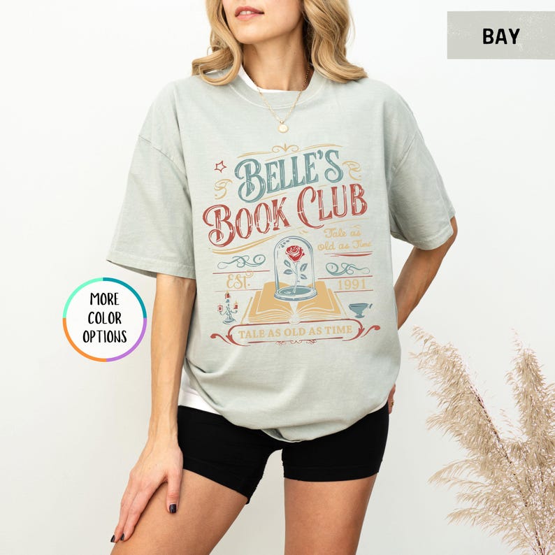 Pu&ograve; includere: T-shirt verde chiaro con la scritta "Belle's Book Club" in rosso e blu. Il design include una rosa in una cupola di vetro, un libro aperto e la frase "Tale as old as time".