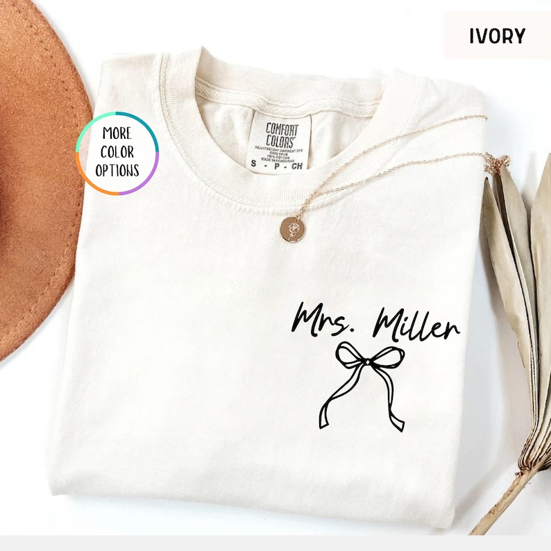 Puede incluir: Camiseta de color marfil con el texto "Mrs. Miller" y un lazo. La camiseta tiene cuello redondo y mangas cortas. Un collar dorado con un colgante est&aacute; drapeado sobre la camiseta. Las palabras "More Color Options" est&aacute;n en un c&iacute;rculo a la izquierda.