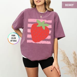 May include: A berry-colored short-sleeve t-shirt featuring a graphic of a red strawberry with green leaves centered over horizontal light pink brushstroke stripes. The text BERRY appears in the top right corner and MORE COLOR OPTIONS is on the left.
