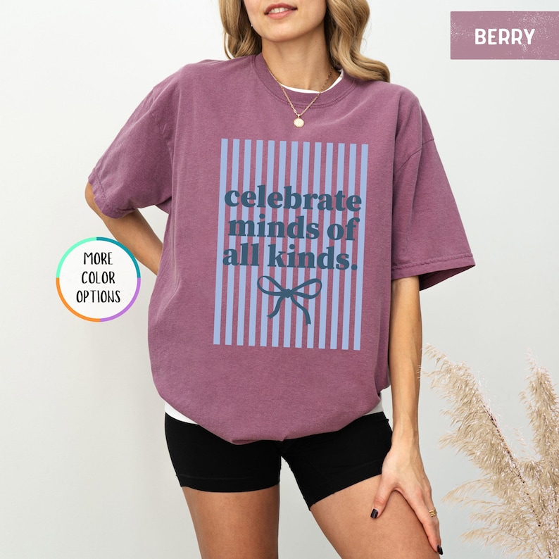Puede incluir: Camiseta color bayas con el texto "celebrate minds of all kinds" en un dise&ntilde;o de rayas azules y blancas. La camiseta tiene un corte holgado y mangas cortas. La modelo lleva shorts negros.