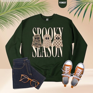 May include: A dark green long-sleeve sweatshirt featuring the text "SPOOKY SEASON" in cream lettering. Three ghost illustrations are centered, each with a different pattern: camouflage, leopard print, and a brown and cream checkerboard design.