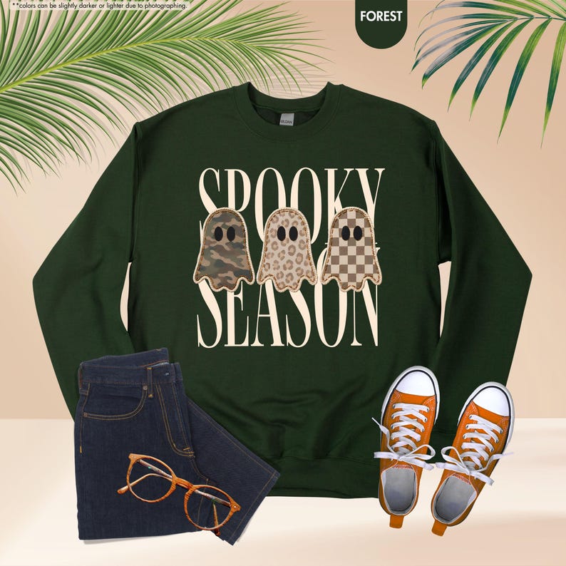 May include: A dark green long-sleeve sweatshirt featuring the text "SPOOKY SEASON" in cream lettering. Three ghost illustrations are centered, each with a different pattern: camouflage, leopard print, and a brown and cream checkerboard design.