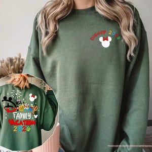 Puede incluir: Una sudadera de cuello redondo color verde militar con un peque&ntilde;o icono blanco de la cabeza de Mickey Mouse con un lazo rojo y el texto "DISNEY 2026" en el pecho. El dise&ntilde;o trasero muestra personajes de dibujos animados coloridos, fuegos artificiales y el texto "DISNEY FAMILY VACATION 2026".