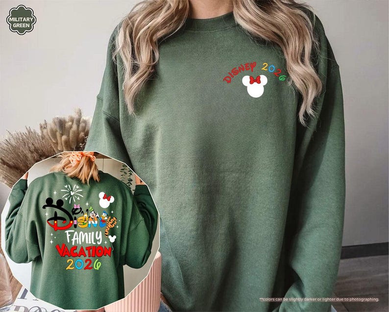 Puede incluir: Una sudadera de cuello redondo color verde militar con un peque&ntilde;o icono blanco de la cabeza de Mickey Mouse con un lazo rojo y el texto "DISNEY 2026" en el pecho. El dise&ntilde;o trasero muestra personajes de dibujos animados coloridos, fuegos artificiales y el texto "DISNEY FAMILY VACATION 2026".