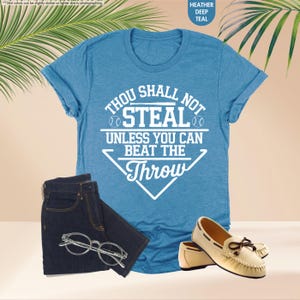 May include: A teal t-shirt with the text "THOU SHALL NOT STEAL UNLESS YOU CAN BEAT THE THROW" in white, paired with dark blue jeans, clear-framed glasses, and cream-colored loafers with brown bows. The shirt is heather deep teal.