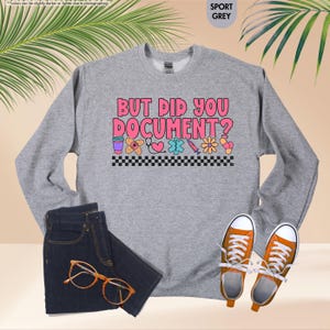 Puede incluir: Sudadera gris deportiva con el texto "BUT DID YOU DOCUMENT?" en rosa y blanco. El dise&ntilde;o incluye gr&aacute;ficos de tem&aacute;tica m&eacute;dica. La imagen tambi&eacute;n muestra unos vaqueros azul oscuro, gafas naranjas y zapatillas naranjas.