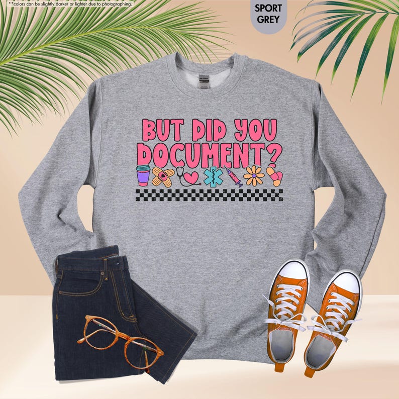 Puede incluir: Sudadera gris deportiva con el texto "BUT DID YOU DOCUMENT?" en rosa y blanco. El dise&ntilde;o incluye gr&aacute;ficos de tem&aacute;tica m&eacute;dica. La imagen tambi&eacute;n muestra unos vaqueros azul oscuro, gafas naranjas y zapatillas naranjas.