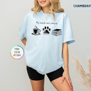 May include: Light blue t-shirt with the text "My needs are simple." The graphic includes a coffee cup, a paw print, and a stack of books. The shirt is a chambray color and has "More Color Options" text.