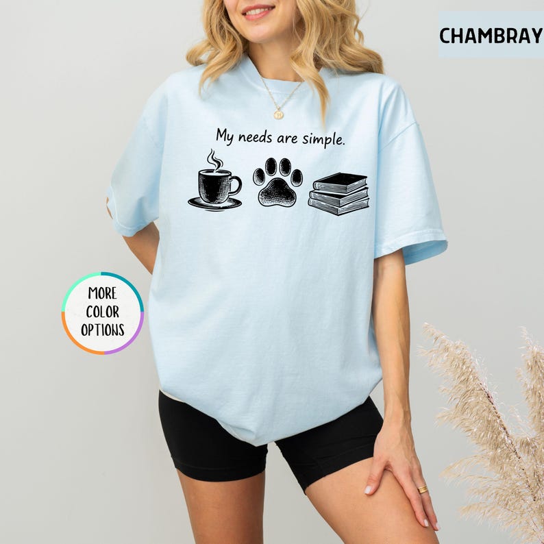 May include: Light blue t-shirt with the text "My needs are simple." The graphic includes a coffee cup, a paw print, and a stack of books. The shirt is a chambray color and has "More Color Options" text.