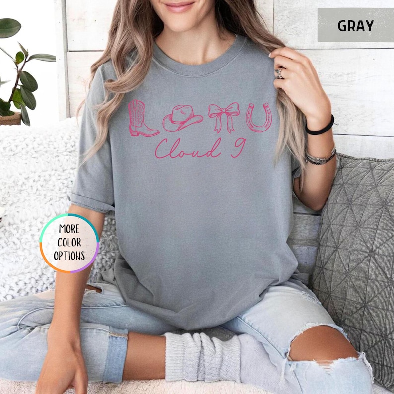 May include: Gray t-shirt with a pink graphic featuring a cowboy boot, hat, bow, and horseshoe, with the words "Cloud 9" in cursive. The shirt is a solid color and has short sleeves.