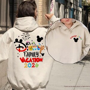 Puede incluir: Una sudadera con capucha color arena que presenta un gr&aacute;fico grande en la espalda con el texto Disney Family Vacation 2026, orejas de Mickey Mouse, fuegos artificiales y personajes. El pecho muestra un peque&ntilde;o icono de Mickey Mouse y Disney 2026.