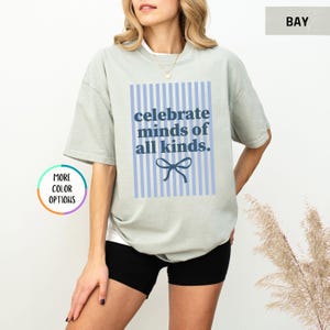 Puede incluir: Camiseta verde claro con el texto azul "celebrate minds of all kinds" sobre un fondo a rayas azules y blancas. Un lazo azul est&aacute; debajo del texto. La camiseta es de corte holgado.