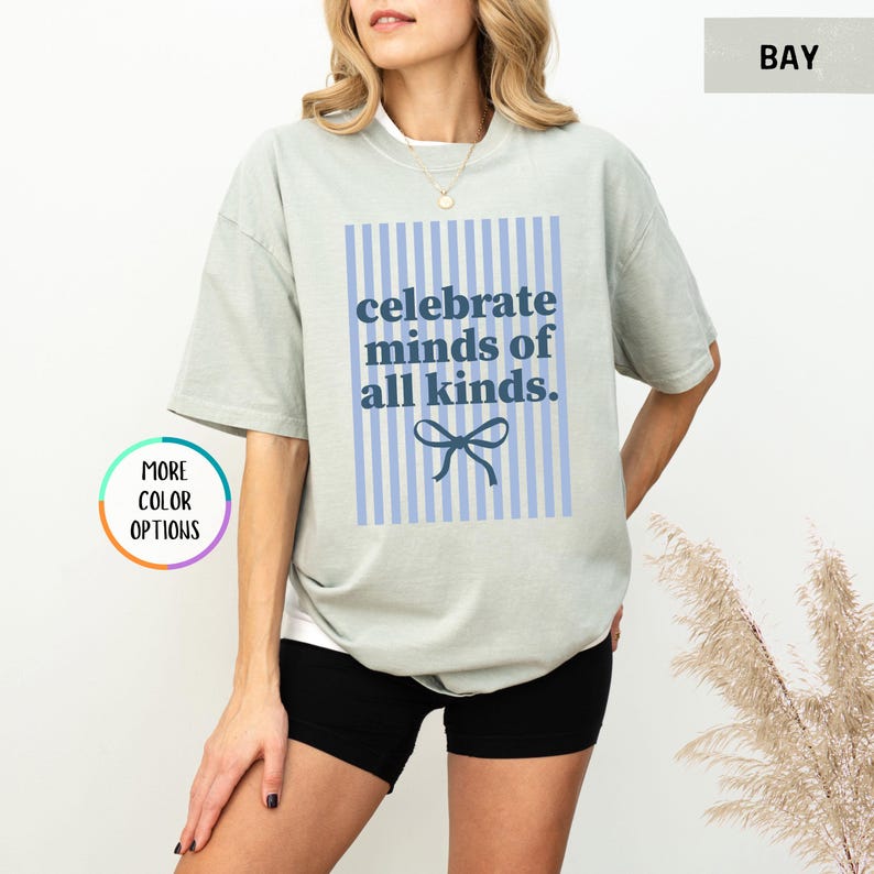 Puede incluir: Camiseta verde claro con el texto azul "celebrate minds of all kinds" sobre un fondo a rayas azules y blancas. Un lazo azul est&aacute; debajo del texto. La camiseta es de corte holgado.
