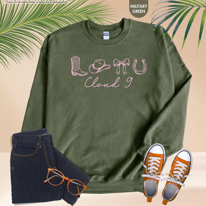 May include: A military green sweatshirt with the text "LOTU Cloud 9" and pink graphics of a cowboy boot, hat, bow, and horseshoe. The image also includes dark blue jeans, orange trainers, and eyeglasses.