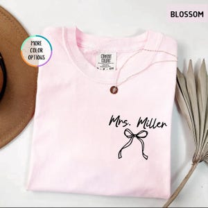 Puede incluir: Una camiseta rosa claro con el texto "Mrs. Miller" y un lazo negro. La camiseta tiene cuello redondo y una etiqueta Comfort Colors. Un collar dorado con un colgante est&aacute; sobre la camiseta. La palabra "BLOSSOM" est&aacute; en la esquina superior derecha.