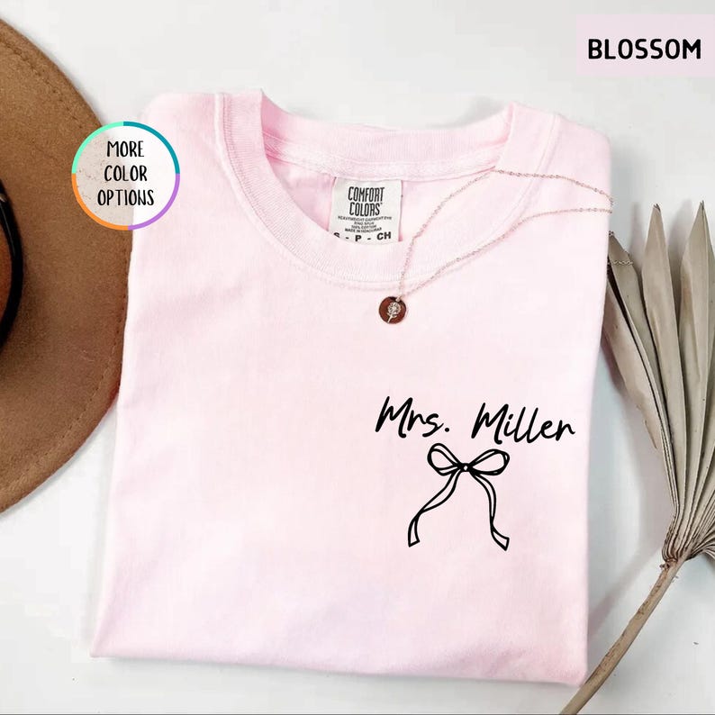 Puede incluir: Una camiseta rosa claro con el texto "Mrs. Miller" y un lazo negro. La camiseta tiene cuello redondo y una etiqueta Comfort Colors. Un collar dorado con un colgante est&aacute; sobre la camiseta. La palabra "BLOSSOM" est&aacute; en la esquina superior derecha.