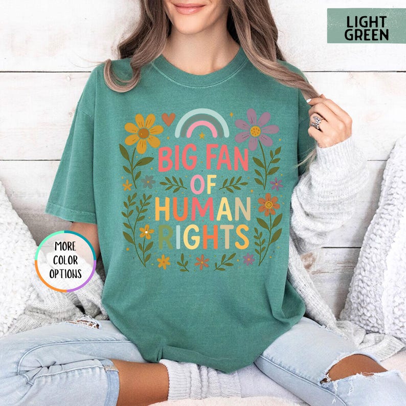 May include: A light green t-shirt with the text "BIG FAN OF HUMAN RIGHTS" in colorful letters. The design includes a rainbow, flowers, and leaves. The shirt is a comfortable, casual style.