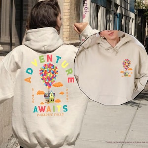Puede incluir: Sudadera con capucha de color arena con un gr&aacute;fico colorido de una casa levantada por globos. El texto "ADVENTURE AWAITS PARADISE FALLS" est&aacute; impreso en la parte posterior. La sudadera tiene un bolsillo delantero y una capucha.
