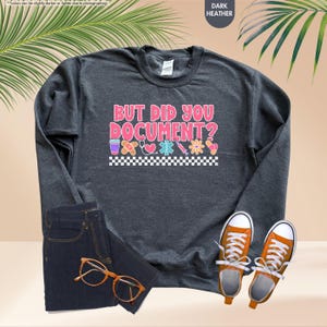 Puede incluir: Sudadera gris oscuro con el texto "BUT DID YOU DOCUMENT?" en rosa y blanco. El dise&ntilde;o incluye gr&aacute;ficos de tem&aacute;tica m&eacute;dica. La imagen tambi&eacute;n muestra unos vaqueros, zapatillas naranjas y gafas.